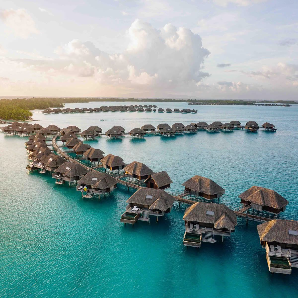 Rows of thatched overwater villas curving across a Bora Bora lagoon at sunset